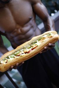 Midsection of shirtless man holding hot dog while standing in gym