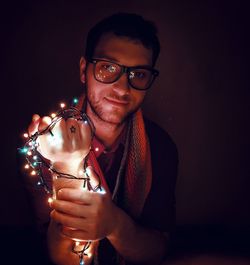 Portrait of young man wearing eyeglasses while holding illuminated string lights in darkroom