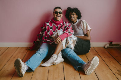 Cheerful hipster woman and gay man sitting on hardwood floor against pink wall at home