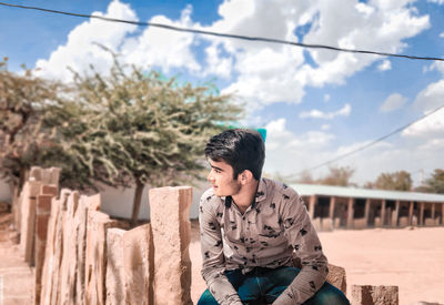 Young man looking away while sitting on tree against sky