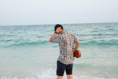 Rear view of young man holding violin while standing at beach against sky during sunset