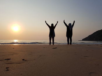 Rear view of friends standing at beach against sky during sunset