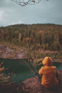 Rear view of woman wearing hood while sitting on rock against river 