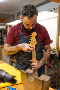 Serious male master in apron standing near table with different equipment while fixing electric guitar in light workshop