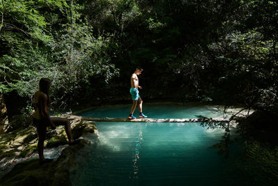 Traveling man walking along log and crossing lake while woman waiting and standing on shore