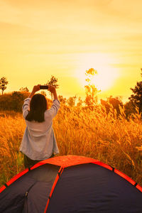 Rear view of woman photographing with mobile phone while standing by tent during sunset