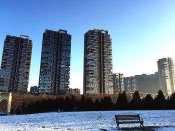 Empty bench on snow against tall buildings