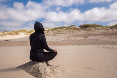 Rear view of person wearing black hooded jacket sitting on rock at beach