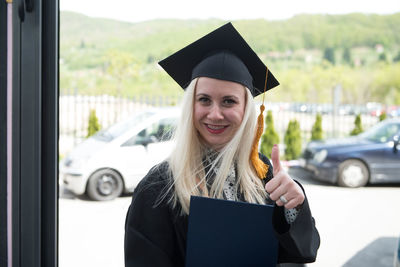 Portrait of smiling young woman wearing graduation gown standing against window