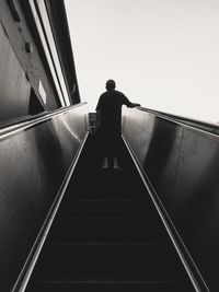 Rear view of man standing on escalator against clear sky