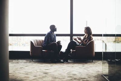 Male and female coworkers discussing while sitting against window at workplace