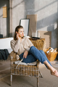 A beautiful young woman in jeans and a jumper sits on a wicker chair by the fireplace at home 