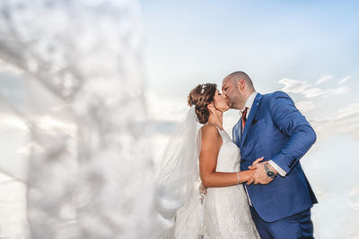 Low angle view of newlywed couple kissing against sky