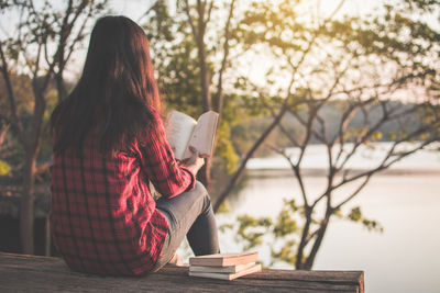 Rear view of woman reading book while sitting by lake