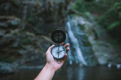 Cropped hand of person holding navigational compass against waterfall