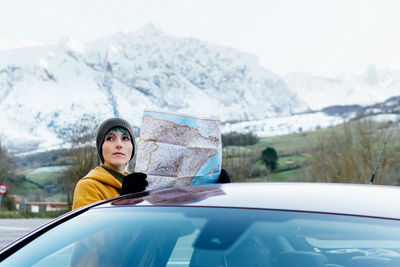 Attractive young female in outerwear standing with territory map near modern car during road trip in snowy mountainous terrain on cold winter day