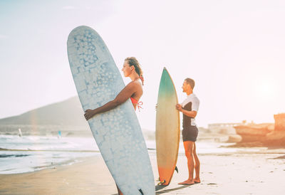 Side view of man and woman with surfboards standing at beach against sky