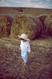 Boy a child in a straw hat and blue pants stands in a mowed field with stacks in the summer