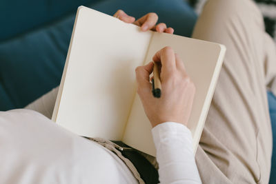 From above faceless lady in white shirt sitting on sofa and writing in notebook working on business project