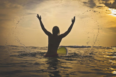 Rear view of playful woman splashing water while sitting on surfboard in sea