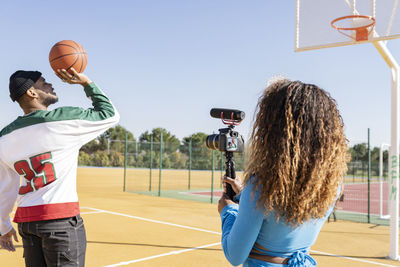 Female filming male friend playing basketball on sports court