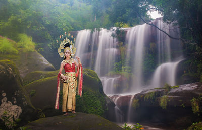 Portrait of beautiful young woman wearing crown and traditional clothing while standing against waterfall in forest
