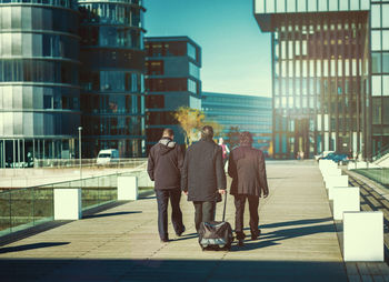 Rear view of businessmen walking on footpath towards modern buildings in city