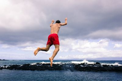 Full length of shirtless man jumping in sea against cloudy sky