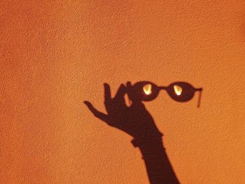 Shadow of hand holding sunglasses on orange wall during sunny day