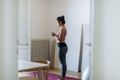 Fit ethnic woman surfing phone standing on yoga mat at home