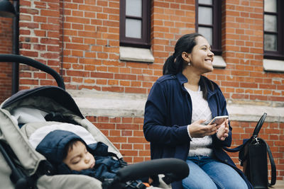 Smiling mother using smart phone while son sitting in stroller against building in city