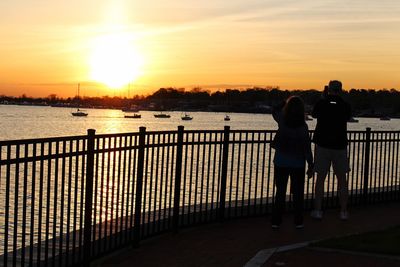 Rear view of people standing by railing against sky during sunset
