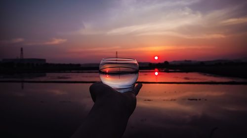 Cropped hand holding drinking glass against sky during sunset