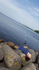 Tilt shot of boy fishing while lying on rocks in lake against sky