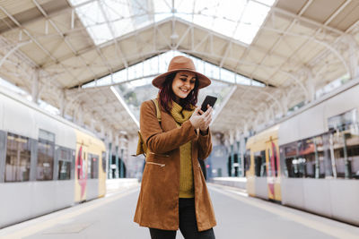 Smiling woman using smart phone while standing at railroad station platform