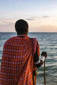 Rear view of man standing at beach against sky during sunset