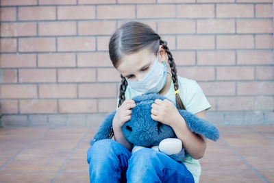 Girl holding a toy while sitting against brick wall