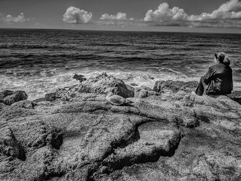 Rear view of men sitting on rock by sea against sky