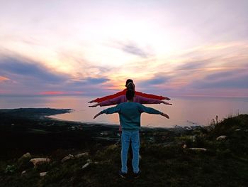 Rear view of woman with arms raised standing on field against sky during sunset