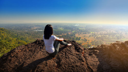 A  woman is sitting on an unusual rock on a mountain overlooking forest