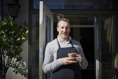 Portrait of smiling owner holding coffee mug while standing outside store