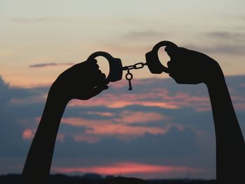 Silhouette cropped hands holding handcuffs against cloudy sky during sunset