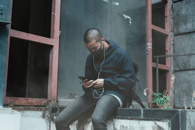 Young man wearing mask listening music while sitting outdoors