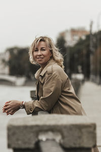 A middle-aged blonde woman with curly hair stands on the river embankment in the city.