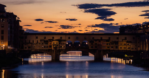 Arch bridge over river amidst buildings against sky during sunset