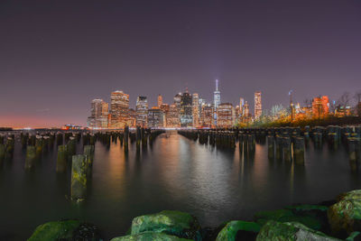 Wooden posts on hudson river by illuminated city skyline against clear sky at dusk