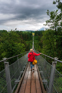 Rear view of woman walking on footbridge in forest against sky