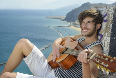 Smiling man playing guitar while sitting on rock