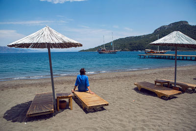 Rear view man in a blue shirt sitting on a sunbed by the beach under an umbrella on the sea