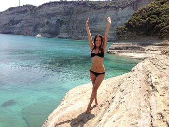Full length of happy sensuous woman in bikini with arms raised standing on rock by sea